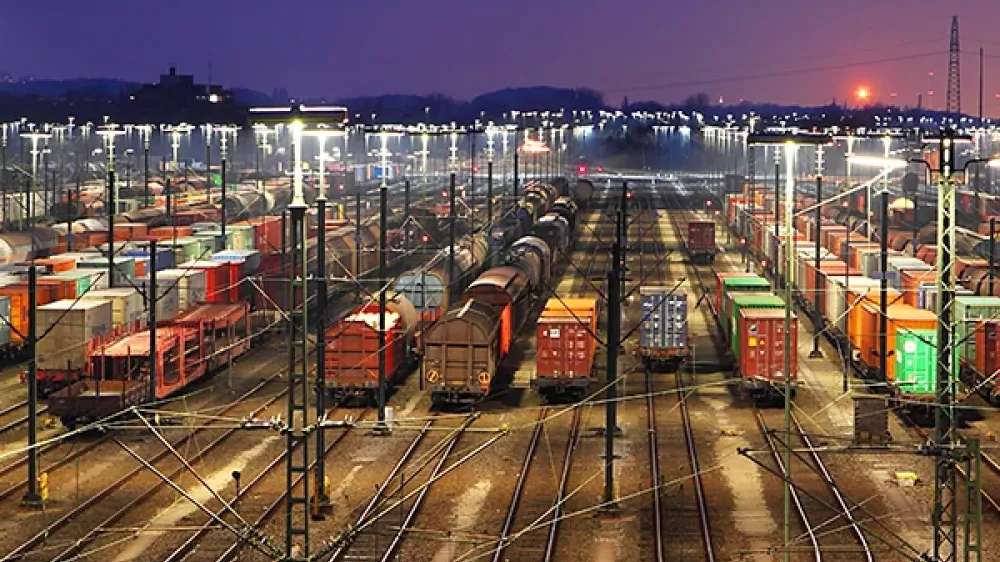 The railway station has many tracks, points and power lines in front of the silhouette of a city.